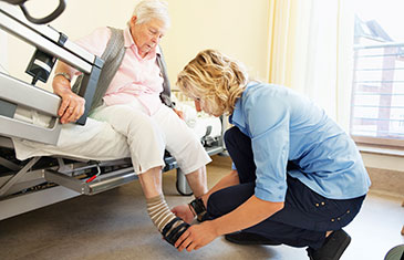 Pflegerin hilft auf Bett sitzender alter Frau in ihre Schuhe