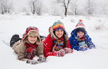 Drei Kinder liegen auf dem Bauch im Schnee mit aufgestützten Händen