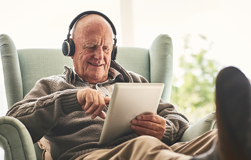 Älterer Mann sitzt im Sessel mit Tablet und Kopfhörern
