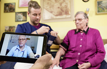 Foto einer Videosprechstunde. Der Arzt ist über ein Tablet per Video zu sehen und eine ältere Dame sitzt mit einem Pfleger auf ihrem Bett zu Hause.