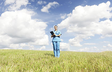 Eine Pflegerin in blauer Krankenhauskleidung steht mit einem Stethoskop um den Hals und einer Mappe auf einem Wiesenfeld.