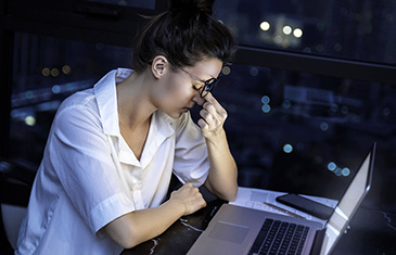 Eine junge Frau, die nachts vor dem Laptop sitzt, massiert sich erschöpft unter ihrer Brille die Augen.