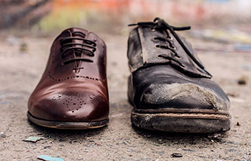 Foto von einem edlen, neuen braunen Schuh neben einem verschmutzten, alten Schuh gleicher Farbe auf sandigem Untergrund