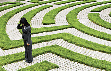 Foto einer Frau im Business-Look. Sie steht in einem Labyrinth und fasst sich an den Kopf. 