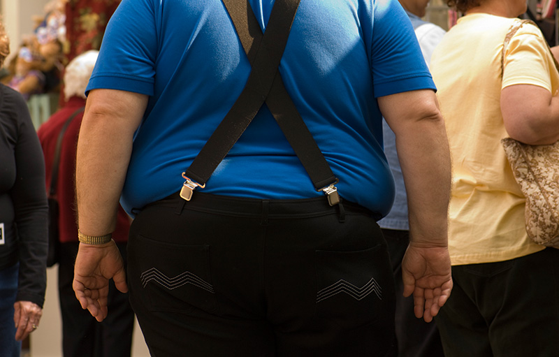 Foto von dickem Mann von hinten im blauen T-Shirt mit scharzer Hose und Hosenträgern