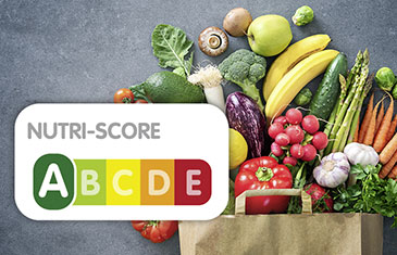 Abbildung des fünffarbigen Nährwertlogos Nutri-Score mit den Buchstaben A bis E. Im Hintergrund: Eine Papptüte mit Obst und Gemüse gefüllt