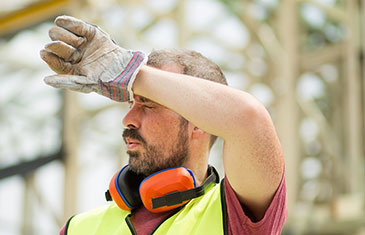 Foto von schwitzendem Arbeiter im roten T-Shirt mit neongelber Weste, Arbeitshandschuhen und Lärmschutzkopfhörern um den Hals, der sich den Schweiß auf der Stirn abwischt.