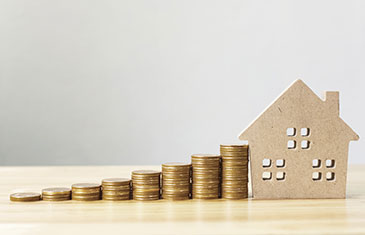 Symbolbild von der Silhouette eines Hauses aus Holz mit einem Geldstapel aus Münzen links daneben, der Richtung Haus ansteigt