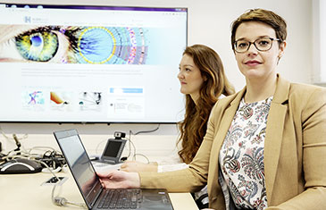 Foto von Medizininformatikerin Antje Wulff (vorne) und ihrer Kollegin Alina Rehberg vor einem großen Bildschirm an der Wand mit der Darstellung eines Auges