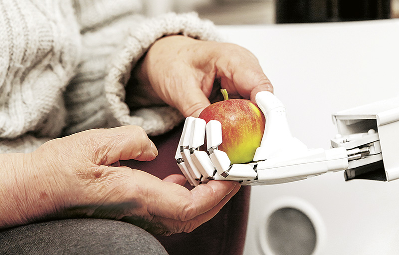 Foto einer Roboterhand, die einem alten Menschen, von dem man nur die Hand sieht, einen Apfel reicht