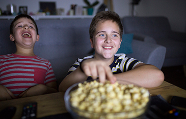 Foto von zwei fröhlichen Jungs auf dem Sofa vor dem Fernseher mit einer großen Schale Süßigkeiten im Vordergrund