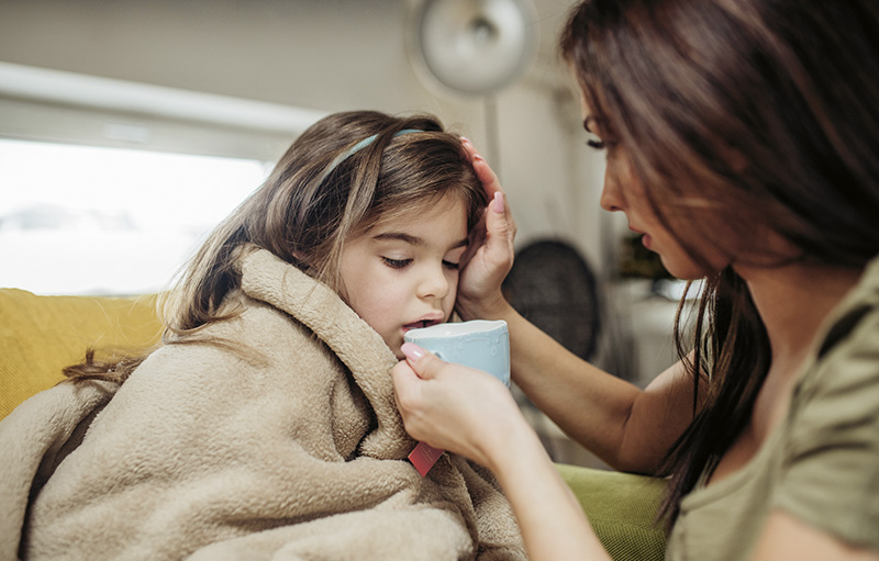 Foto einer jungen Mutter, die einem kranken Mädchen auf dem Sofa, das in eine Decke gehüllt ist, eine Tasse Tee an die Lippen hält