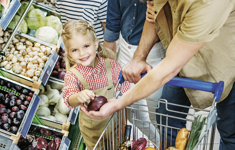 Foto eines kleinen, blonden Mädchens, das eine rote Zwiebel in einen Einkaufswagen legt und zusammen mit zwei Erwachsenen einkauft.