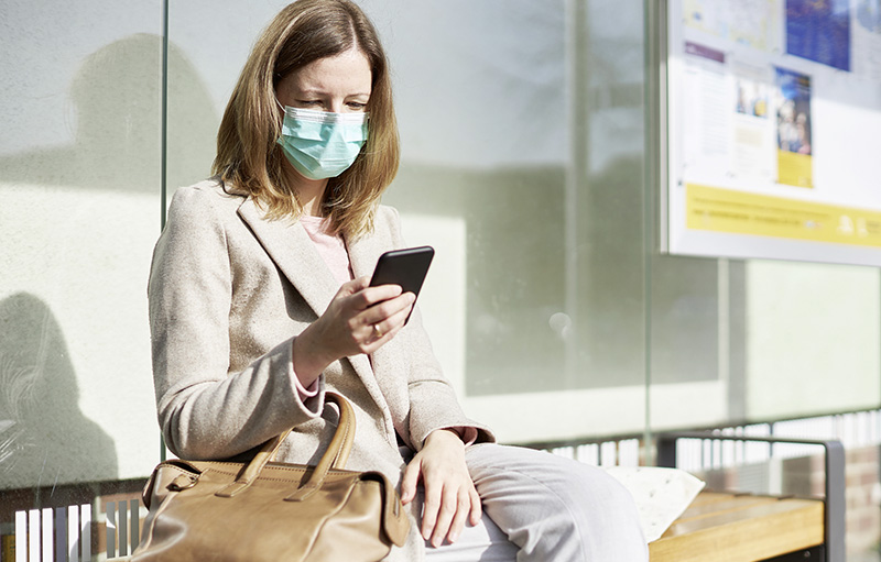 Foto einer Frau im Business-Outfit mit Maske, die in einem öffentlichen Gebäude auf ihr Smartphone blickt