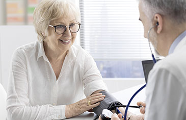 Foto einer älteren Patientin mit Brille, der ein Arzt mit grauem Vollbart den Blutdruck misst