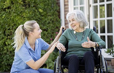 Foto einer Altenpflegerin, die im Freien auf Augenhöhe mit einer alten Dame mit Rollstuhl spricht