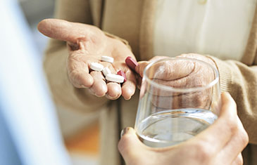 Foto von Händen einer älteren Frau mit Tabletten, jemand reicht ihr ein Glas Wasser dazu
