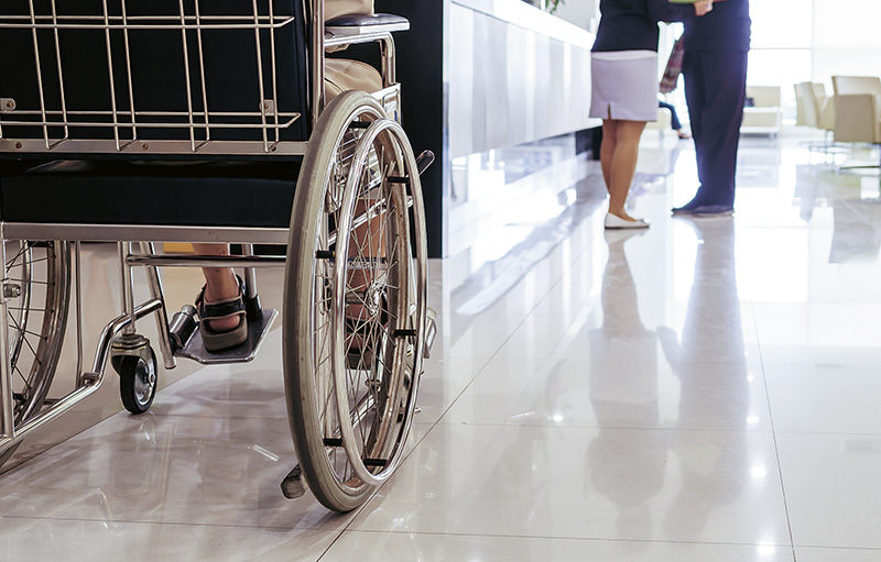 Foto eines Rollstuhls mit einem Patienten. Der Rollstuhl steht auf einem Flur, während weiter entfernt zwei Menschen reden.
