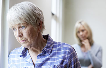 Foto einer traurig blickenden alten Frau im Vordergrund. Im Hintergrund eine nachdenkliche Angehörige.
