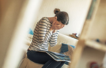 Foto einer sitzenden Frau mit Unterlagen auf dem Schoß, die erschöpft den Kopf in die Hände legt