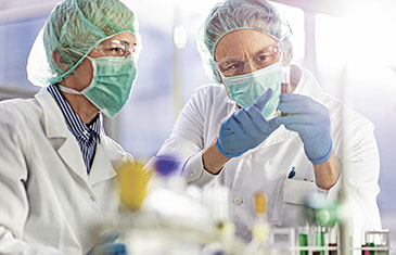 Foto von zwei Männern mit Maske, Haube und Laborkittel im Gespräch über ein Präparat in einem Laborröhrchen
