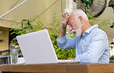 Foto eines alten Mannes mit Vollbart am Laptop, der sich erschöpft an die Stirn fasst.
