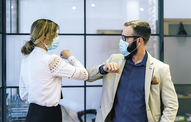 Foto: Eine Frau und ein Mann tragen jeweils einen Mund-Nasen-Schutz und begrüßen sich mit Ellbogen statt Händeschütteln.