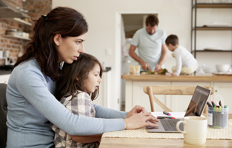 Foto einer vierköpfigen Familie zuhause: Die Mutter sitzt mit Kindergartenkind am Laptop. Der Vater kocht im Hintergrund mit dem Sohn.