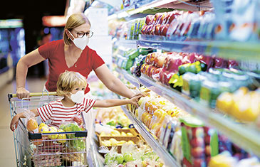 Foto einer Mutter mit Maske beim Einkauf im Supermarkt. Im Einkaufswagen sitzt ein Kleinkind, das ebenfalls eine Maske trägt.