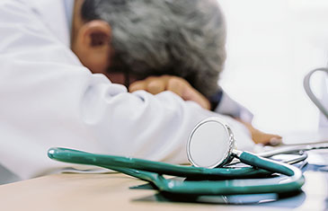Foto eines erschöpften Arztes, der seinen Kopf auf dem Tisch abgelegt hat, vor ihm liegt ein Stethoskop