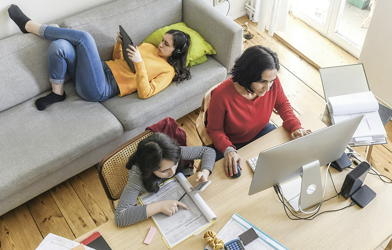 Foto einer Mutter, die am Esstisch mit dem Laptop arbeitet, während eine Tochter auf dem Sofa ins Tablet blickt und die andere neben ihr am Tisch Hausaufgaben macht