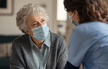 Ältere Frau mit Mundschutz im zugewandten Gespräch mit jünger Pflegekraft