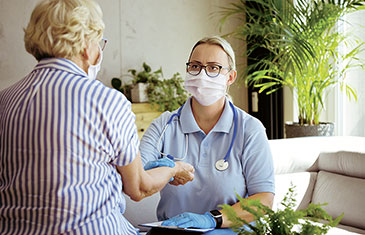Foto einer Pflegerin mit Maske, die bei einer alten Frau - ebenfalls mit Maske - den Blutdruck misst