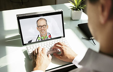 Foto einer Videosprechstunde: im Video zu sehen ist ein Arzt vor weißem Hintergrund, davor sitzt ein Patient an der Tastatur