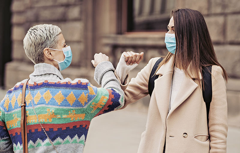Foto von zwei Frauen mit Maske, die sich im Freien mit dem Ellbogen begrüßen