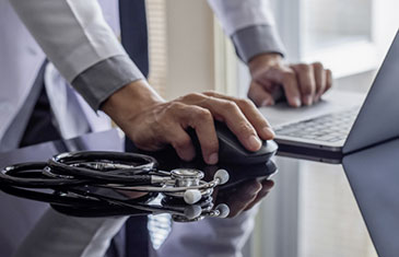 Foto eines Arztes, der stehend an einem Laptop arbeitet, daneben liegt ein Stethoskop