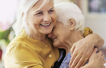 Foto einer mittelalten, grauhaarigen Frau, die eine ältere Frau im Arm hält