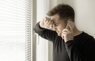 Foto eines Mannes, der mit verzweifelter Miene und geschlossenen Augen mit Handy am Ohr am Fenster steht