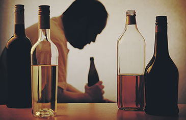 Foto von Flaschen mit Alkohol, im Hintergrund ein Trinker mt Flasche, das Gesicht ist nicht zu erkennen