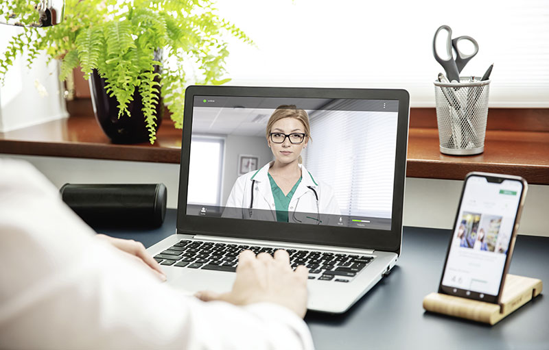 Foto einer Ärztin auf einem Laptop-Bildschirm, vor dem eine Person am Schreibtisch sitzt