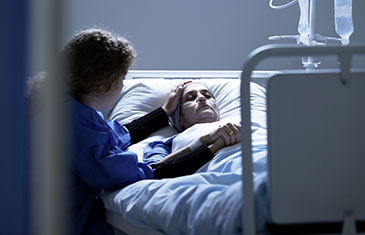 Pflegerin hält im Bett liegender Frau im Krankenzimmer die Hand und legt ihr die andere mitfühlend auf die Stirn