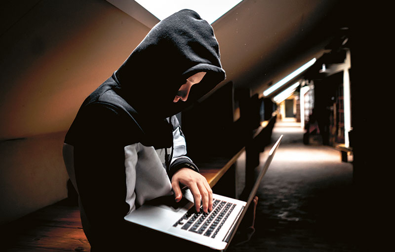 Foto eines Mannes, der unter einer Dachschräge in einen schwarzen Kapuzenpullover gehült am Laptop in seiner Hand tippt