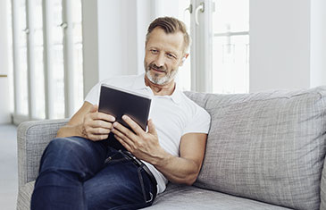 Foto eines älteren Mannes mit Vollbart, der auf dem Sofa sitzt und mit Kopfhörern im Ohr in ein Tablet blickt