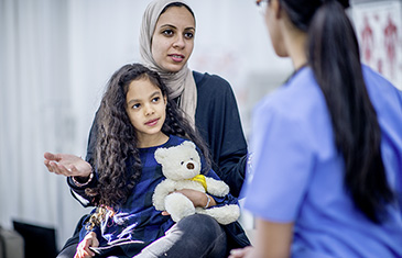 Foto einer jungen muslimischen Frau mit Tochter auf dem Schoß, die mit einer Ärztin im blauen Kittel spricht