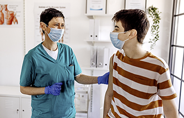 Foto eines Jugendlichen mit Maske im Gespräch mit Medizinerin in grünem Kittel, die ebenfalls eine Maske trägt