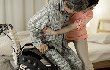 Foto einer Frau mittleren Alters, die einer alten Frau aus dem Rollstuhl hilft, um ins dahinter stehende Bett zu gehen