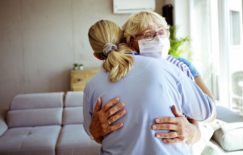 Foto einer Pflegekraft, die eine alte Frau mit Maske umarmt