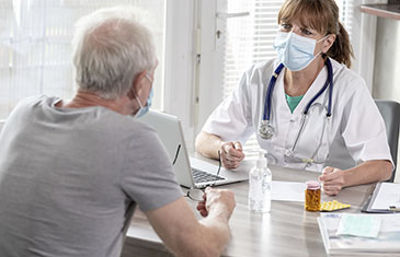 Foto einer Ärztin mit weißem Kittel und Stethoskop, die mit einem älteren Patienten spricht. Beide tragen eine Maske.
