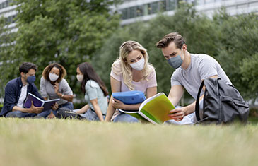 Foto von jungen Studenten und Studentinnen, die in Grüppchen auf einer Wiese sitzen. Alle tragen eine Maske.