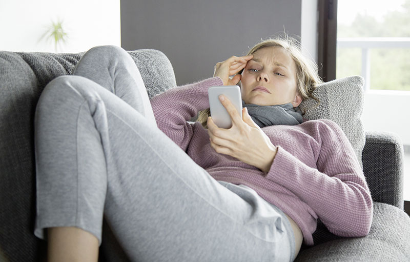 Foto einer jungen Frau auf dem Sofa, die - sichtlich erkrankt - im Liegen ins Handy blickt